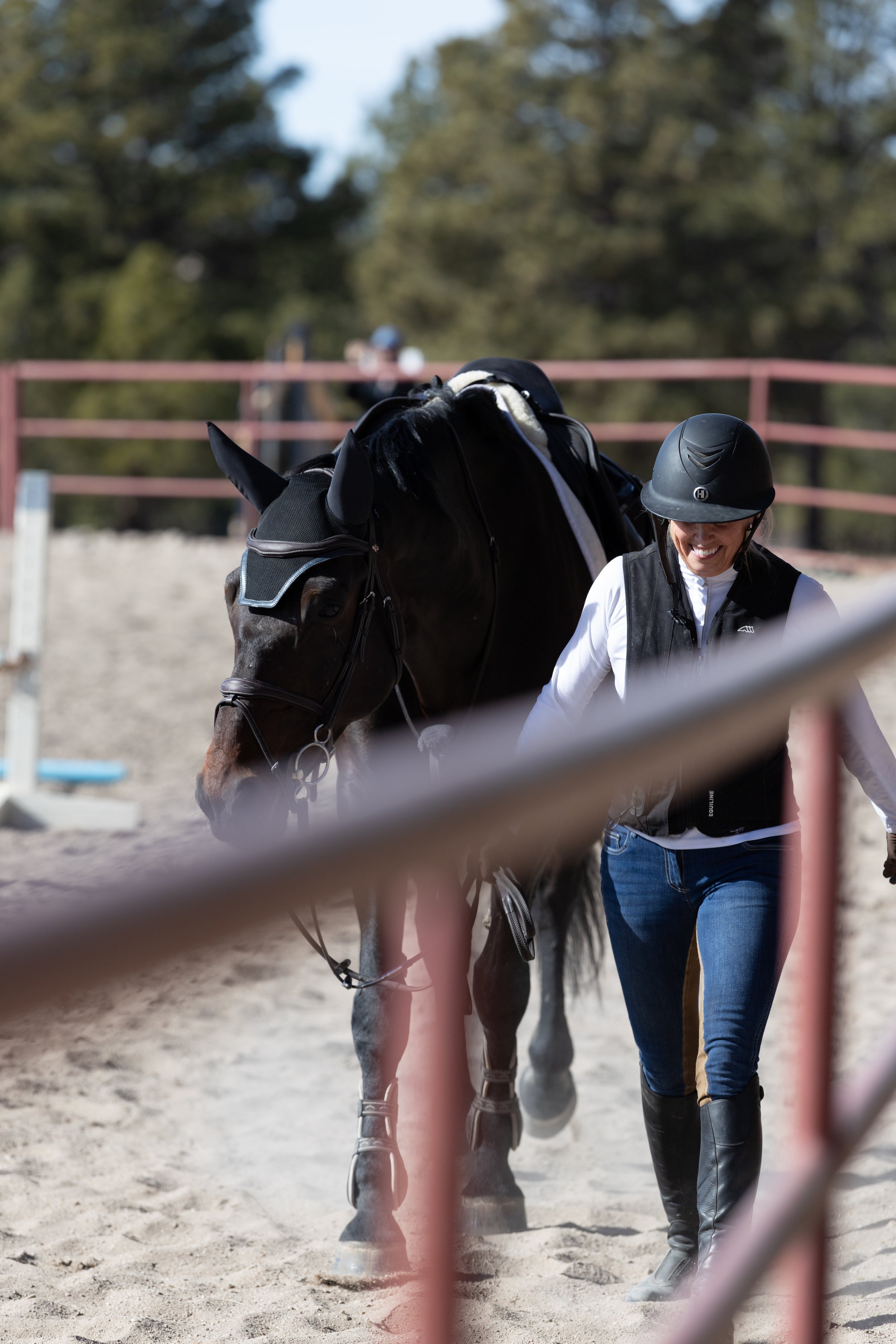 A woman in a riding helmet and vest walking a black horse on a dirt track, with trees in the background.