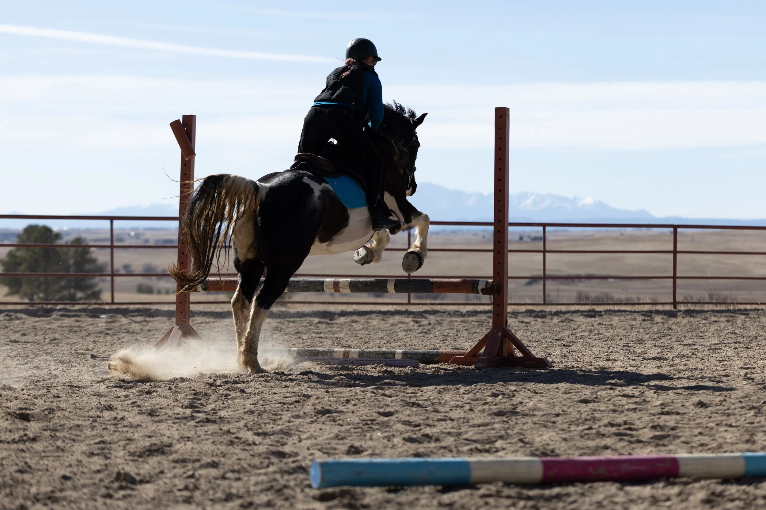 A person riding a black and white horse jumping over a pole in an outdoor equestrian arena.