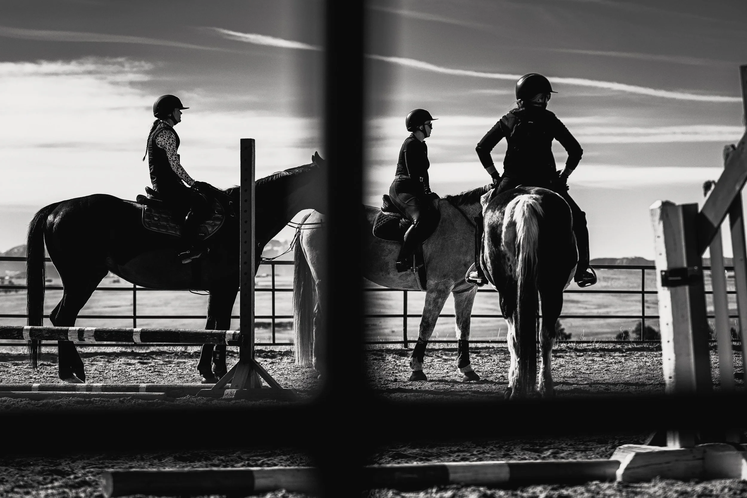 Three people riding horses in an outdoor arena during sunset, view through a fence in black and white.