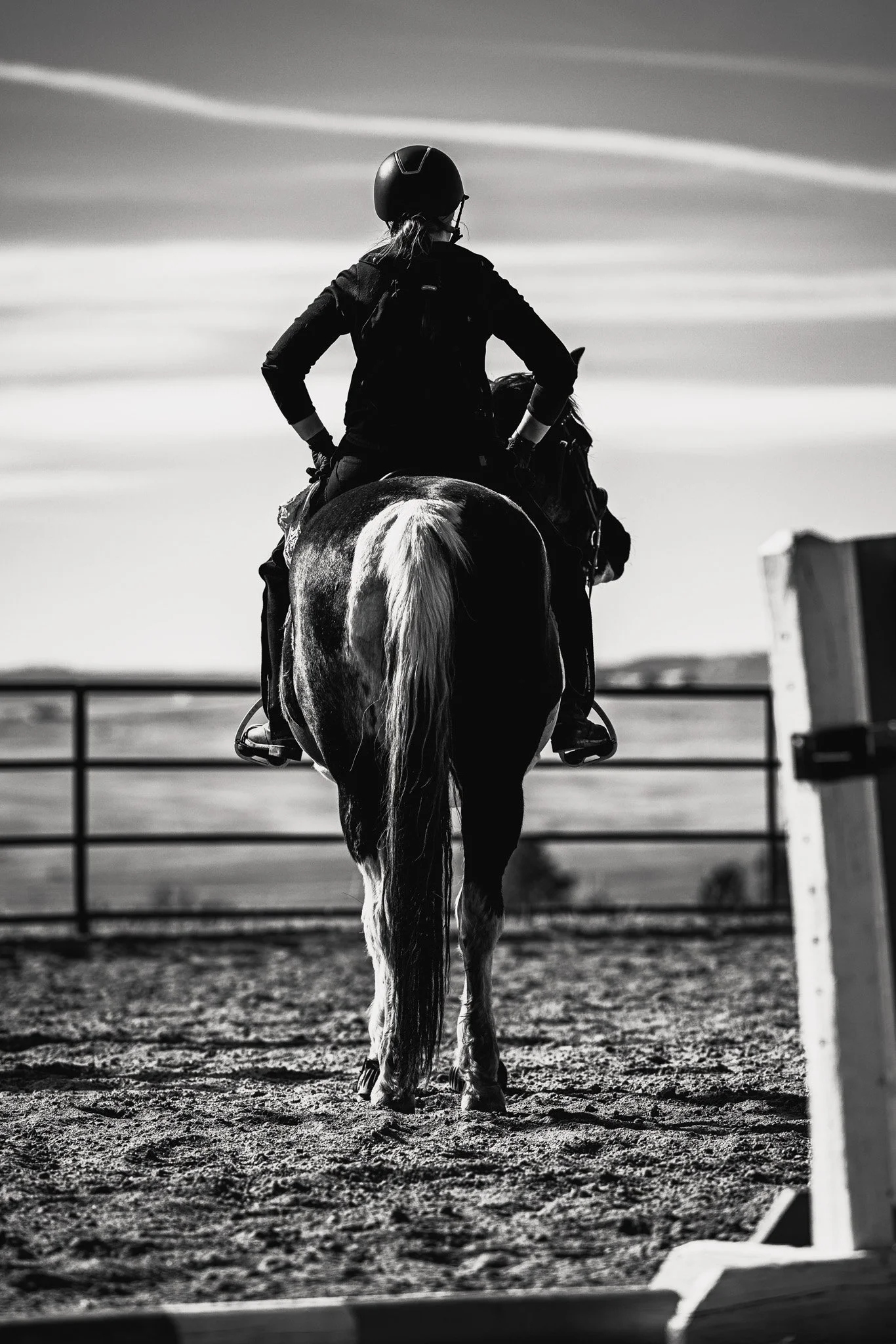 A person riding a horse in an outdoor arena, seen from behind, in black and white.