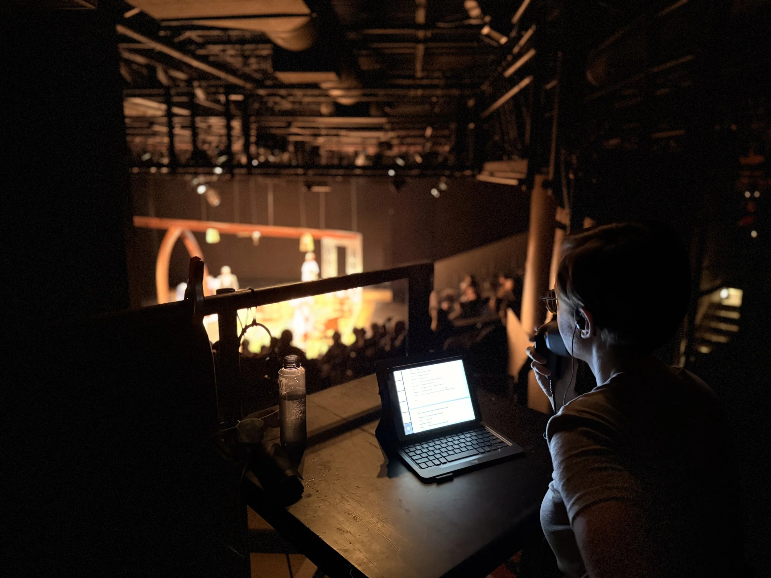 Photo of audio describer from behind. We see their transmitter microphone over their mouth, their laptop in front of them, and an out of focus stage with an audience in front of them