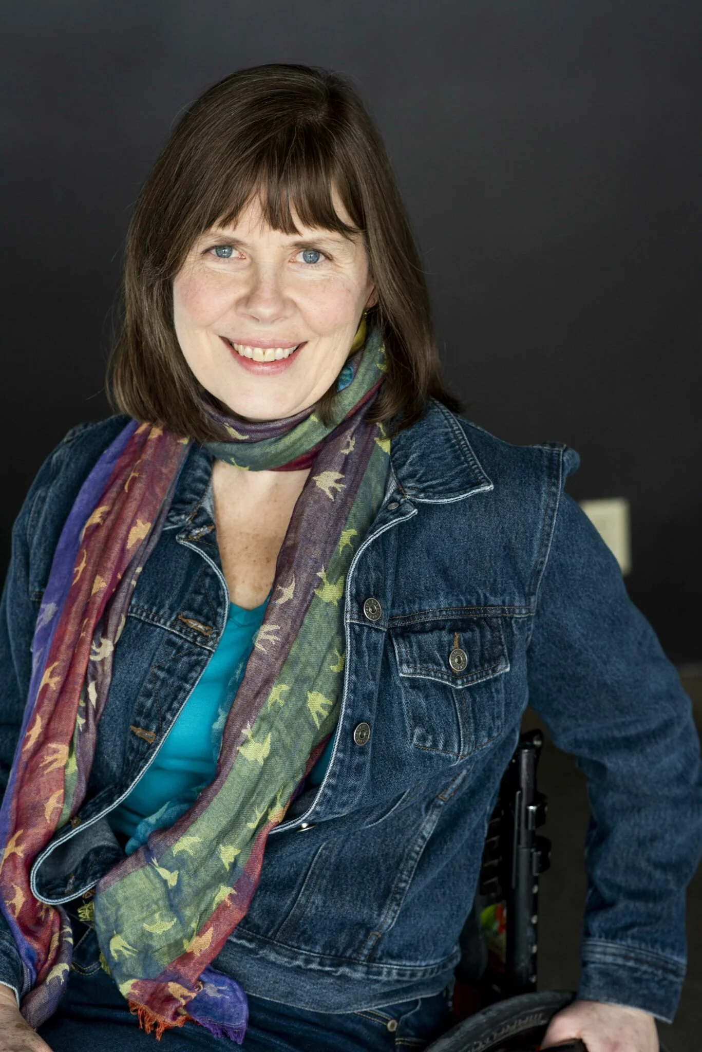 Photo of Debbie Patterson smiling at the camera, sitting in her wheelchair. She has light skin, shoulder length brown hair with bangs, and is wearing a denim jacket, a teal t-shirt, and a colourful linen scarf with a small bird pattern on it