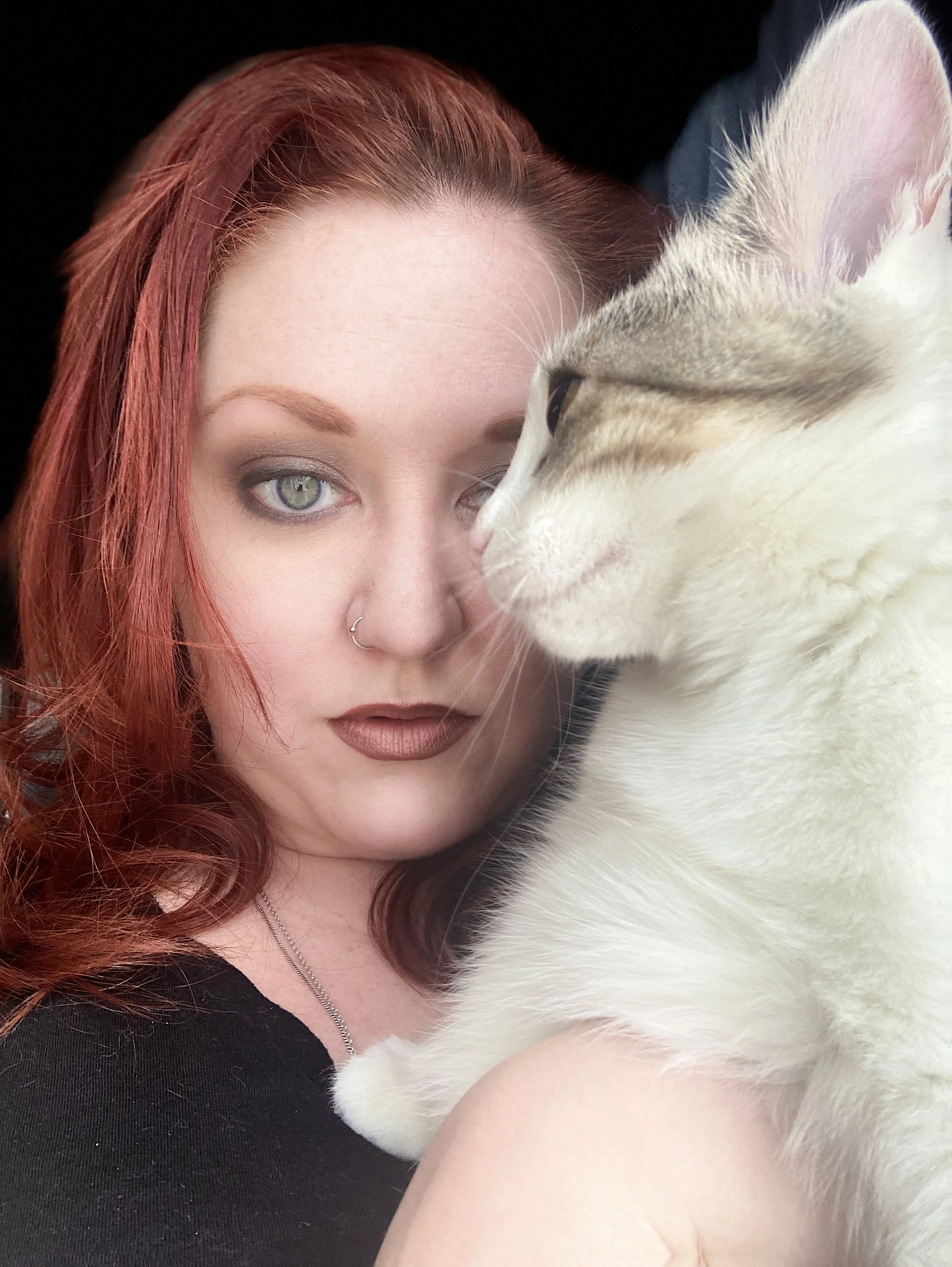 Photo of Theresa Thomson, looking at the camera while holding her cat (on her right, looking to the left). Theresa has light skin, red hair, bright blue eyes, and is wearing a black top.