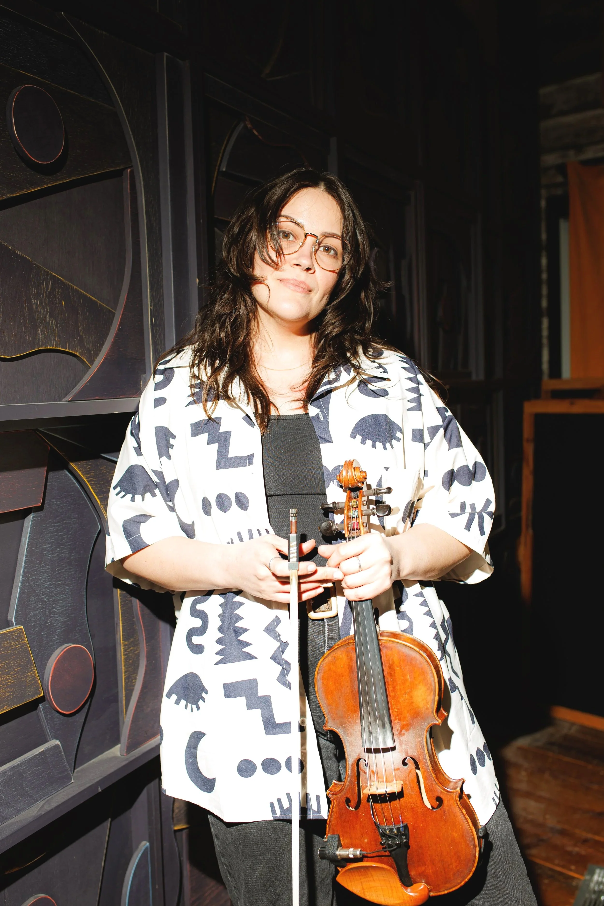 Photo of Sierra Noble. They have light skin, shoulder length brown wavy hair, are wearing round glasses, a black and white geometric patterned top and are holding their fiddle