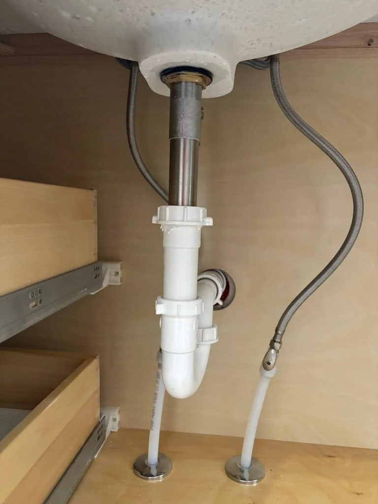 Underneath a kitchen sink showing drainage pipes, flexible water supply lines, and part of a dishwasher connection.