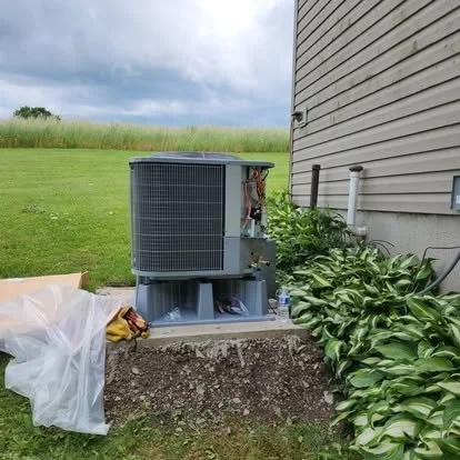 Outdoor air conditioning unit next to house on a concrete slab, with green plants and grass, and a cloudy sky in the background.