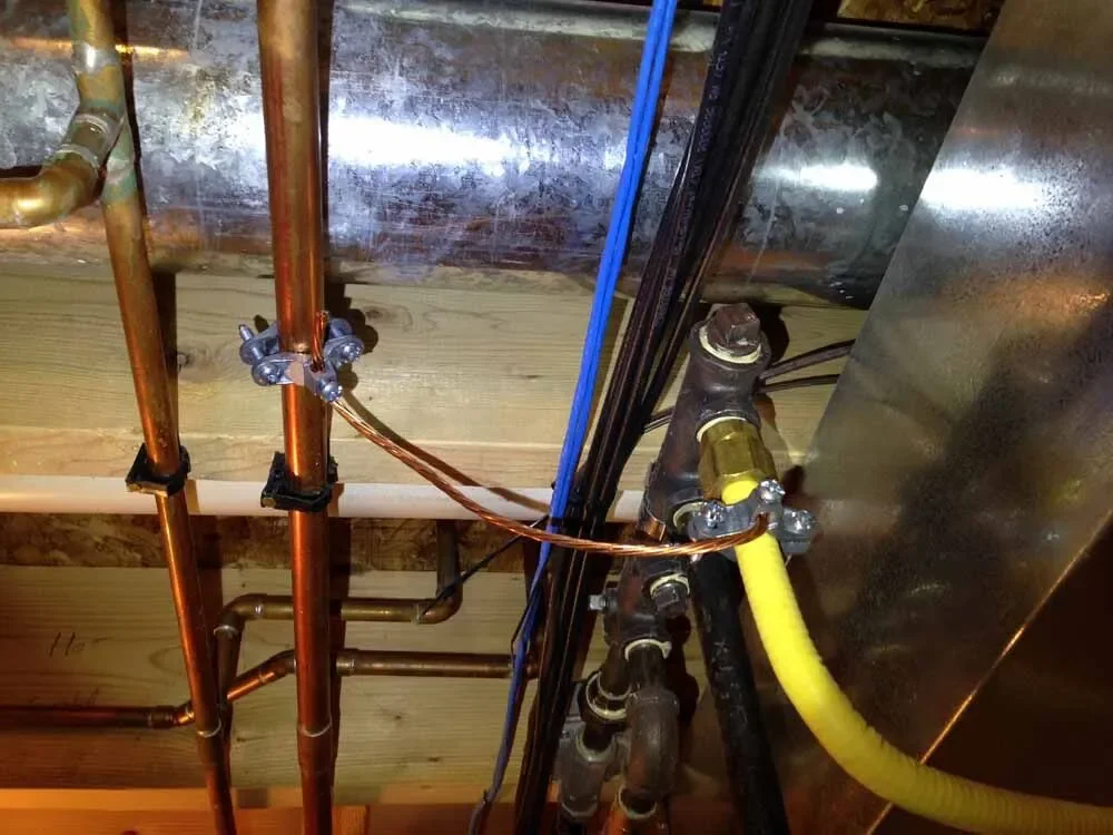 Construction or plumbing setup with copper pipes, a copper tank, black pipes, and a yellow flexible pipe in a building's ceiling.