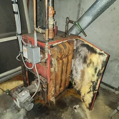 Rusty and burned industrial furnace or boiler with a damaged, blackened side panel in a utility room.