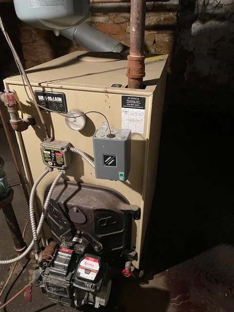 A beige Heil furnace with a black control panel and various pipes and wires connected, located in a basement with brick walls.