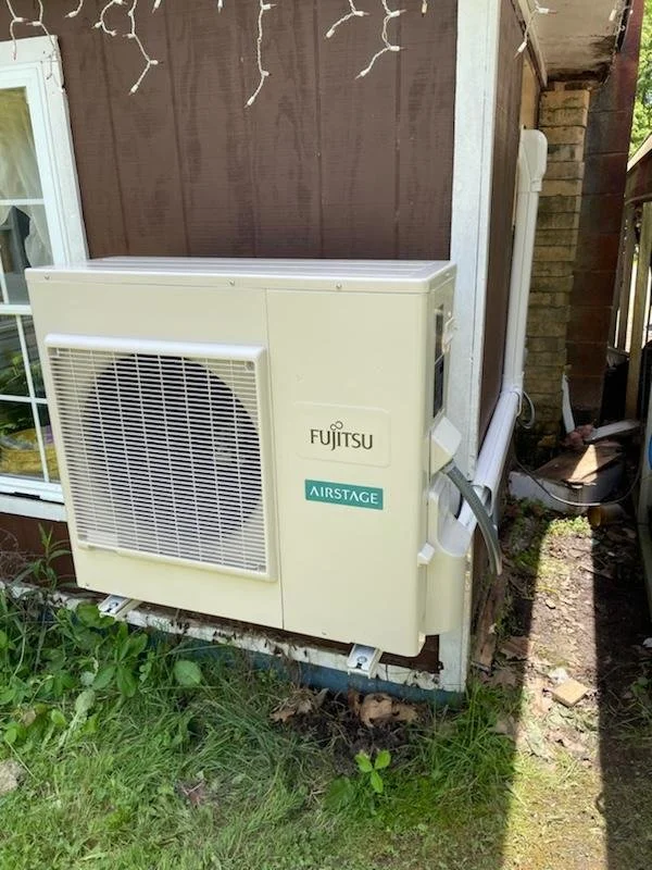 Outdoor Fujitsu AIRSTAGE air conditioning unit installed beside a house, with a window nearby and a small garden area in front.