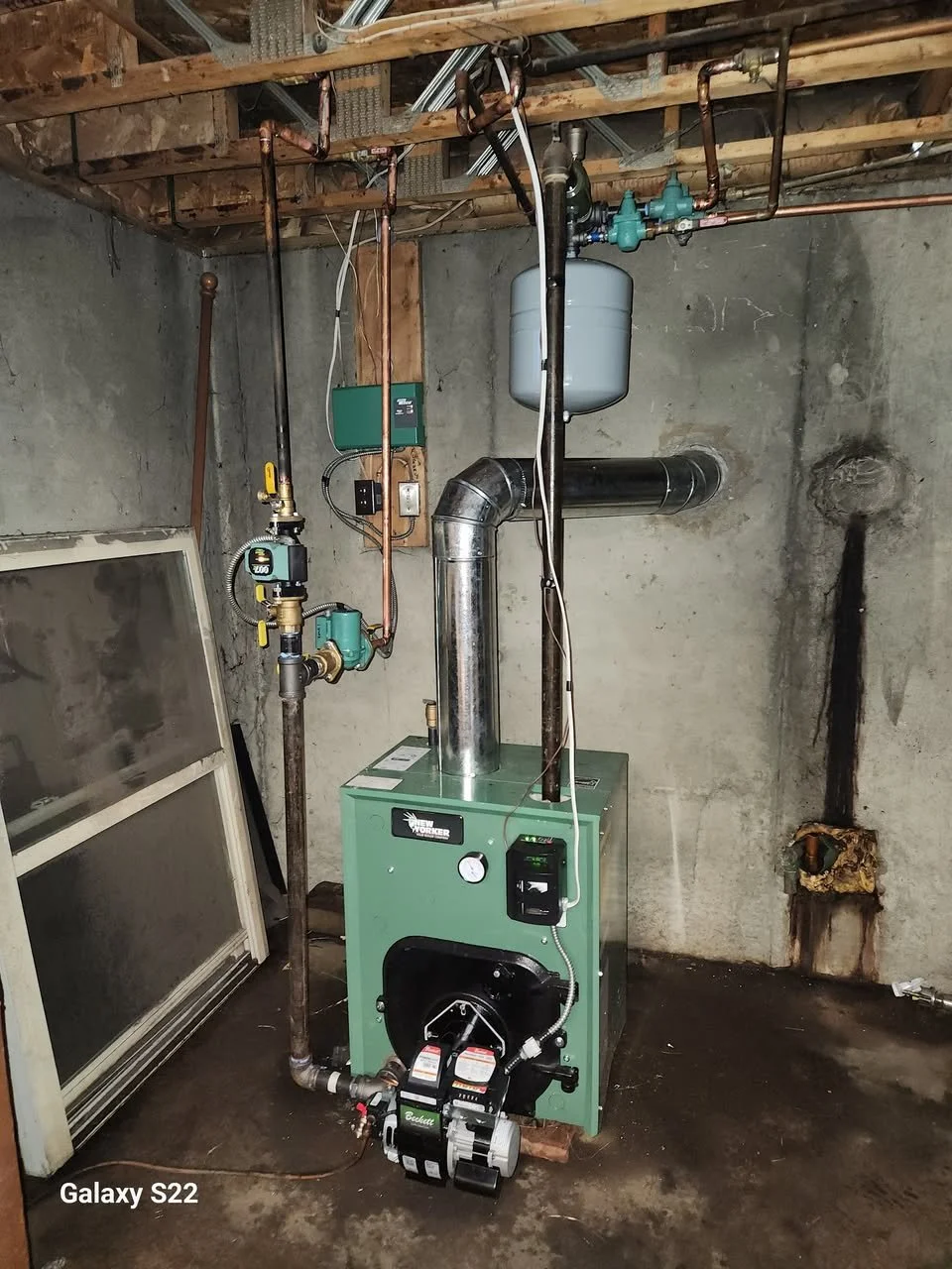 A green furnace or boiler unit installed in a basement with exposed pipes and wall, connected to copper and black metal pipes, with a gauge, a pump, and a ream of wiring visible.