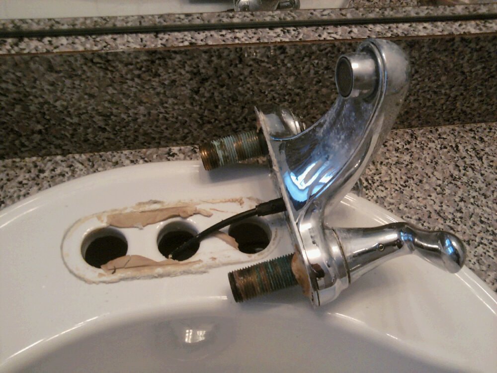 A damaged bathroom sink faucet with two rusty threaded pipes, placed on a countertop, with a hole in the sink and exposed plumbing.