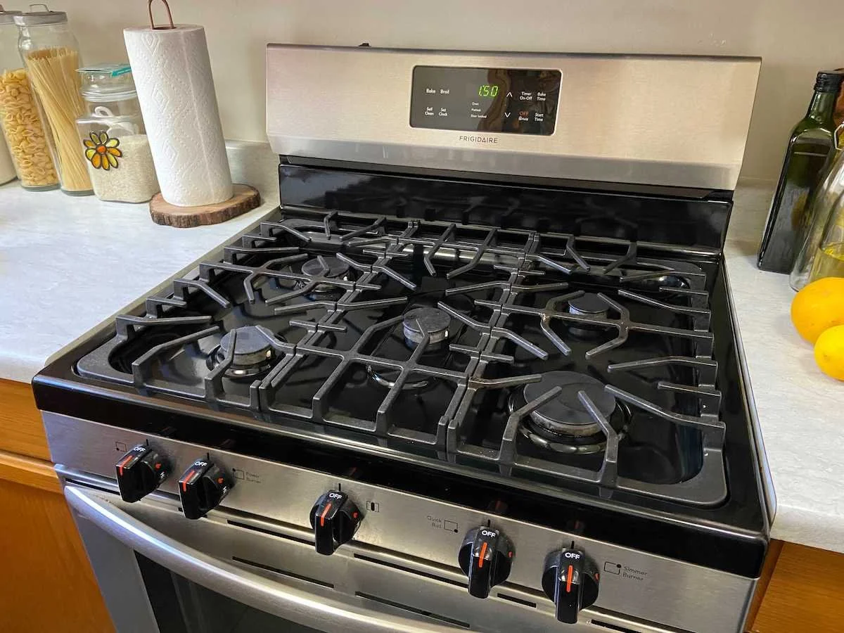A stainless steel gas stove with black grates and control knobs on a kitchen countertop, with a paper towel roll and jars of pasta nearby.