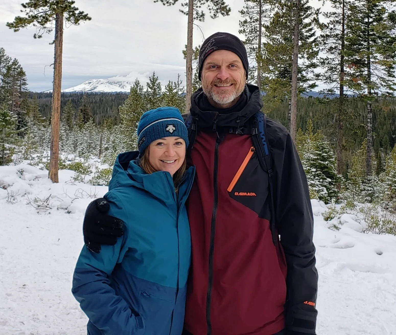 Aly Waibel and James Wood in a snowy forest with pine trees and snow-capped mountains in the background. Both are dressed in winter outdoor clothing.