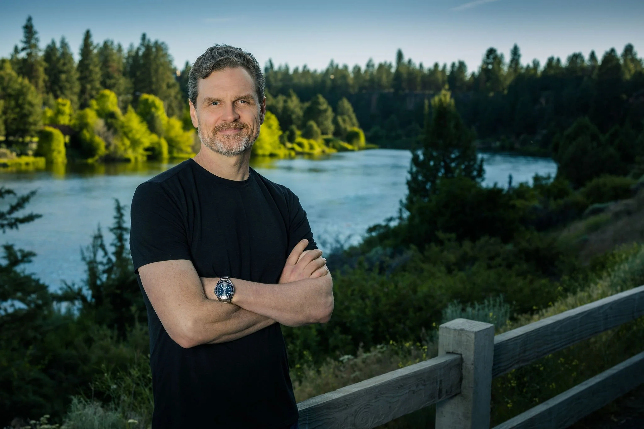 James Wood standing outdoors by a river and lush green trees, wearing a black shirt and a wristwatch, with arms crossed and smiling.