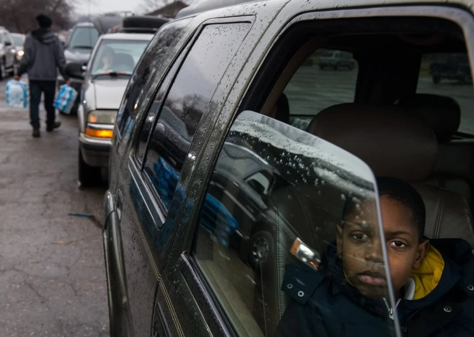A young boy sitting inside a vehicle, looking out the window with reflections of other cars and a person outside in the parking lot. The window has raindrops on it.