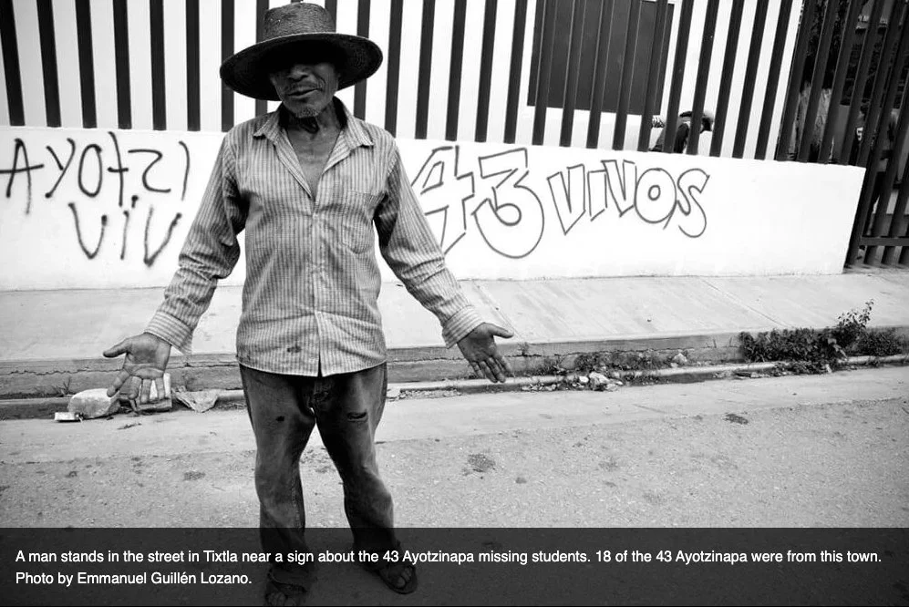 A man stands on the street near a sign about 43 Ayotzinapa students. The sign reads 'Ayotzinapa 43' with the rest obscured. The man is wearing a large hat and striped shirt, with arms outstretched.