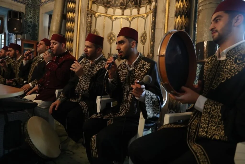 A group of men dressed in traditional Moroccan clothing, some wearing Fez hats, are sitting in a row and performing with microphones and musical instruments in an ornate, decorated room.