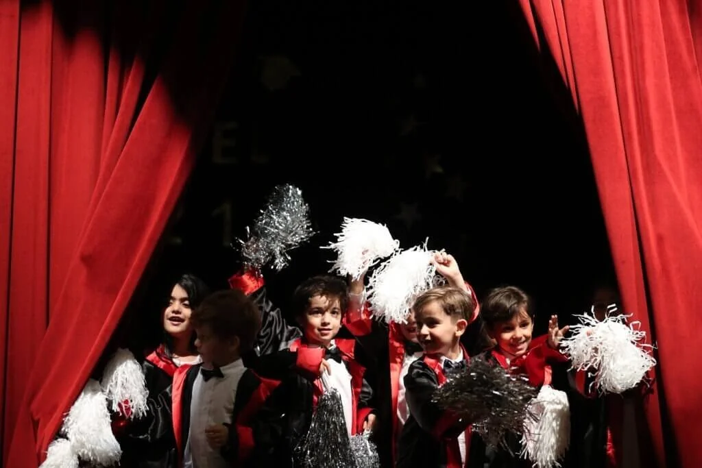 Children dressed in costume with pom-poms on stage behind red curtain.