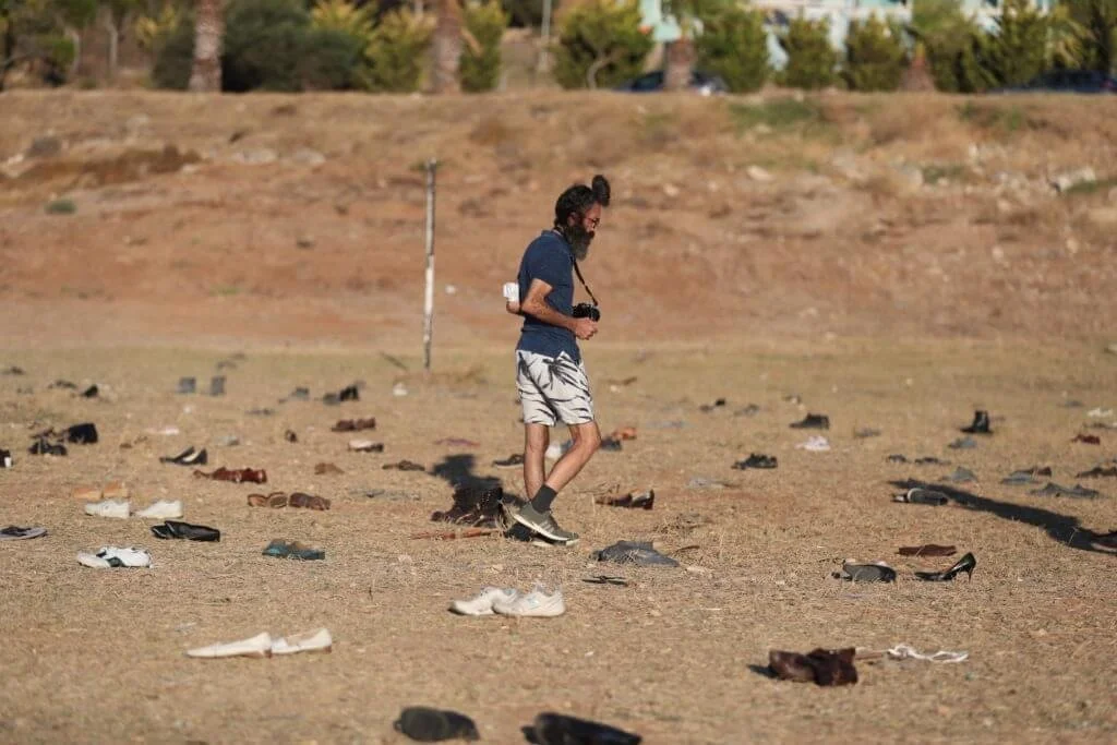 A man with a camera around his neck walking among scattered shoes on a dry field with a hill and trees in the background.