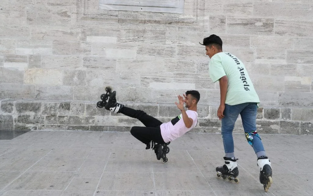 Two young men rollerblading against a stone wall. One is standing and watching, the other is performing a roller skate trick.