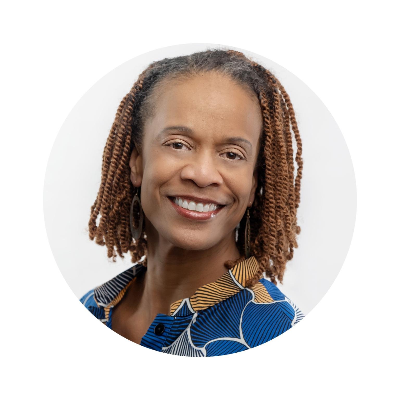 A smiling African American woman with brown, braided hair and wearing a colorful patterned blouse, standing against a white background.