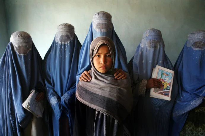 A young girl with a gray hijab and a serious expression standing in front of a group of women wearing blue traditional dresses and head coverings, with one holding a colorful children's book.