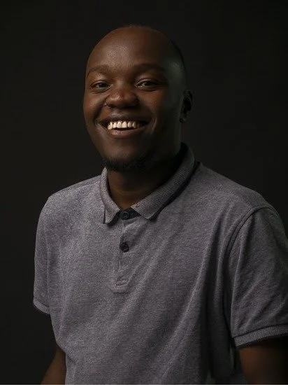 A smiling African man with dark skin, short hair, wearing a gray polo shirt, against a dark background.