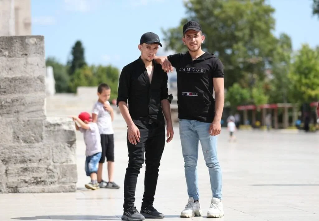 Two young men stand arm-in-arm outdoors on a sunny day, with trees and children in the background.