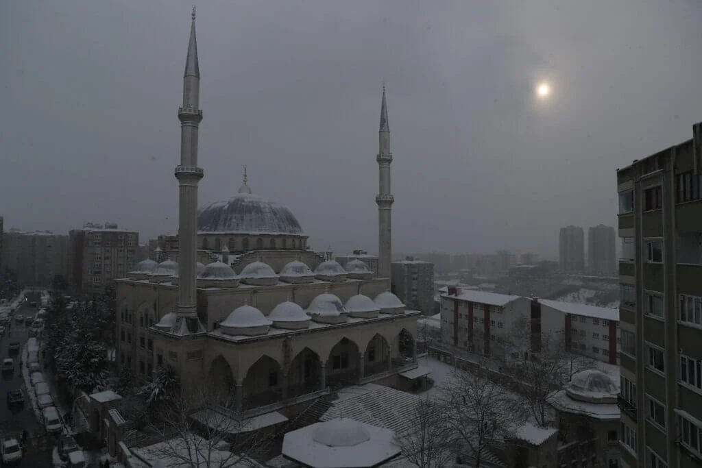 A mosque with snow on its domes and minarets, surrounded by snow-covered buildings and trees under a gray, cloudy sky with the sun faintly visible.