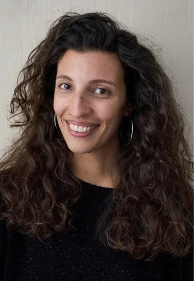 A woman with long, curly brown hair smiling and wearing hoop earrings, standing in front of a light-colored wall.