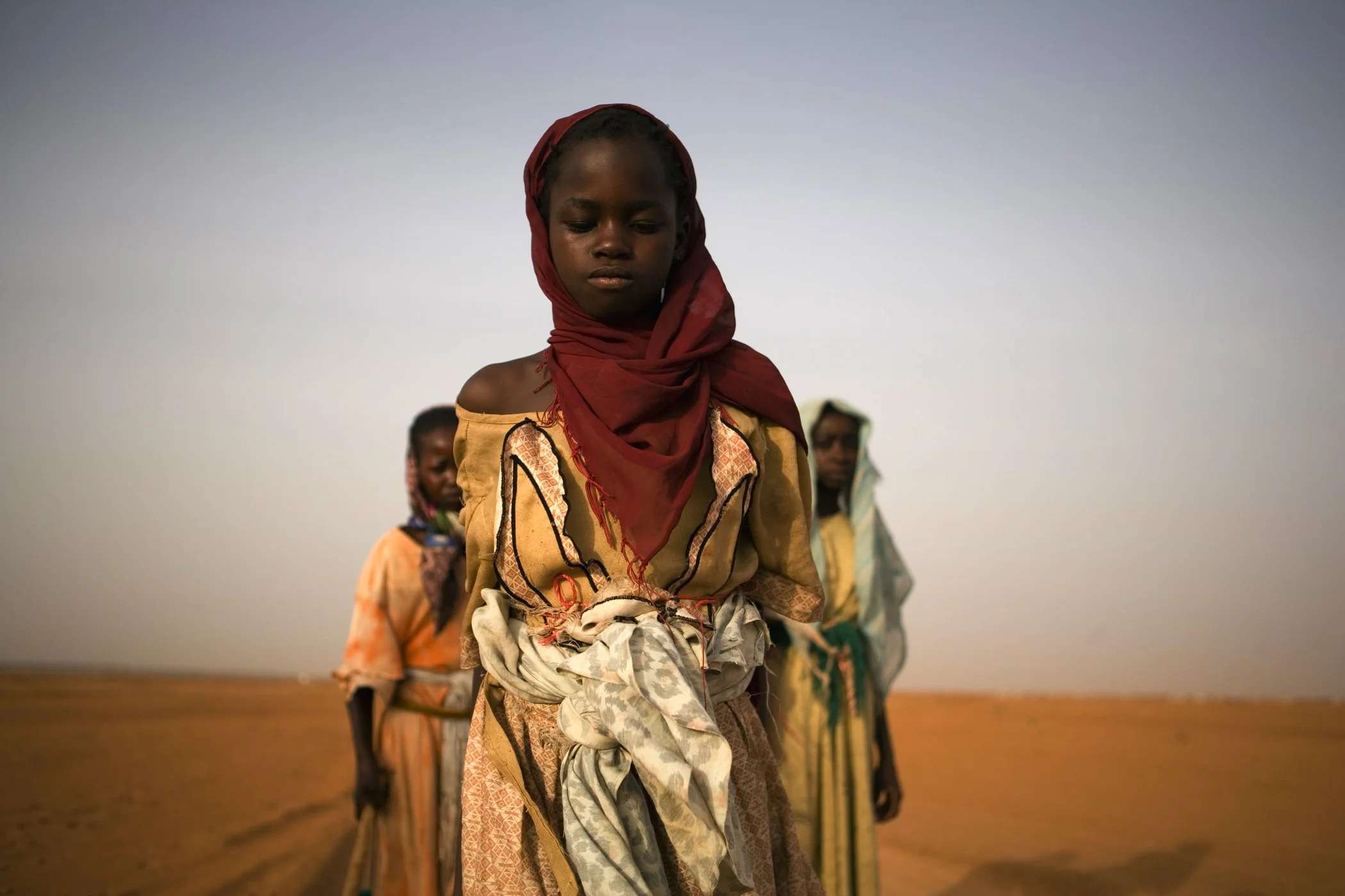 A young girl in traditional dress and headscarf standing outdoors in a desert landscape, with other children in similar attire behind her.