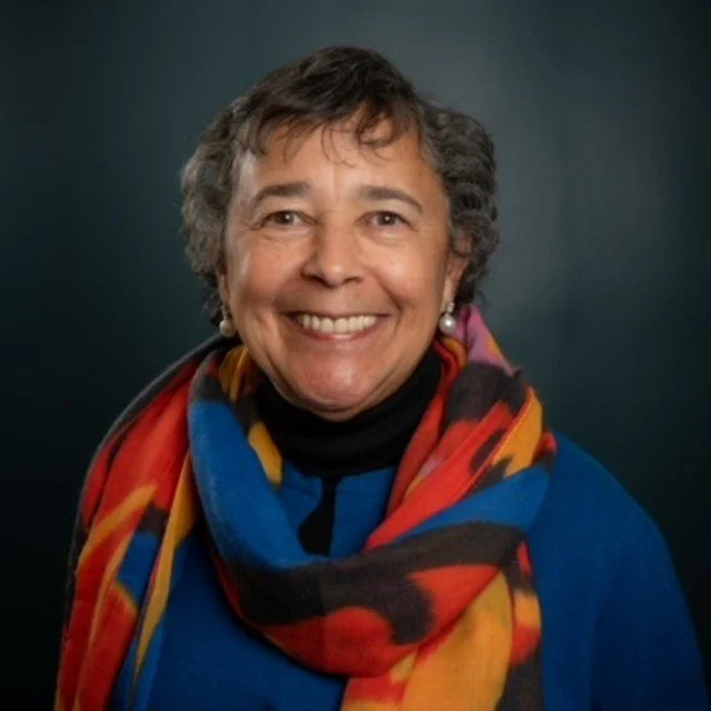 A smiling African American woman with short gray hair wearing a colorful scarf and pearl earrings, posing against a dark background.