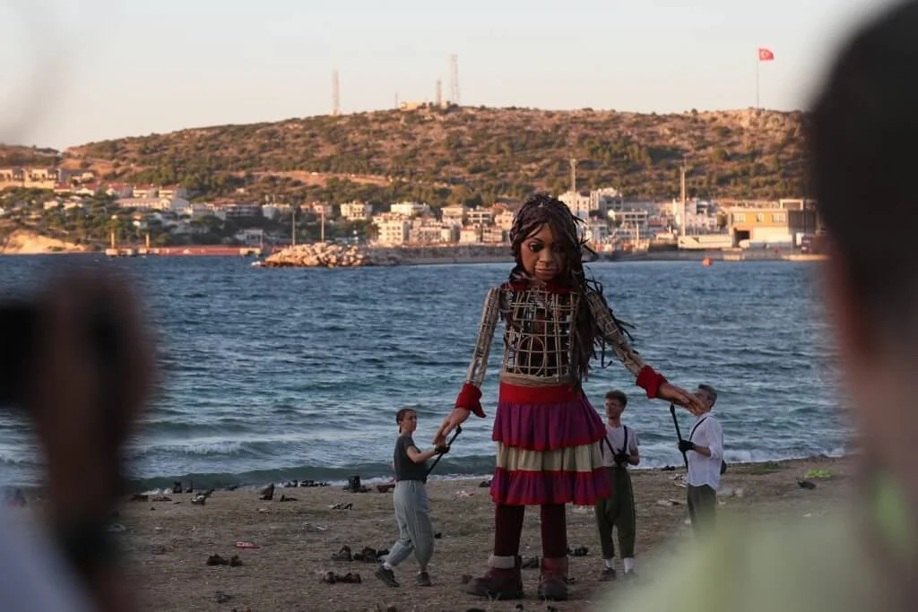 Large puppet of a woman with braids, dressed in a colorful skirt and top, is held up by two people on a beach with water and a cityscape in the background.