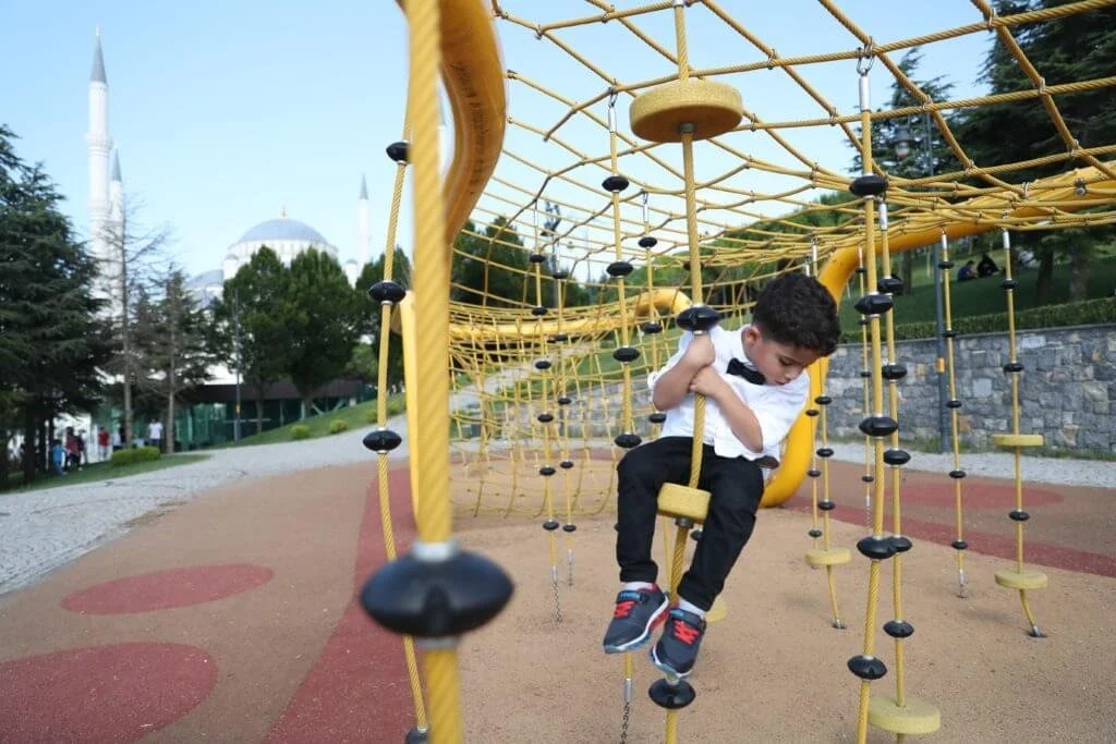Child in a white shirt and black pants climbing on a yellow jungle gym in a park with a mosque in the background.