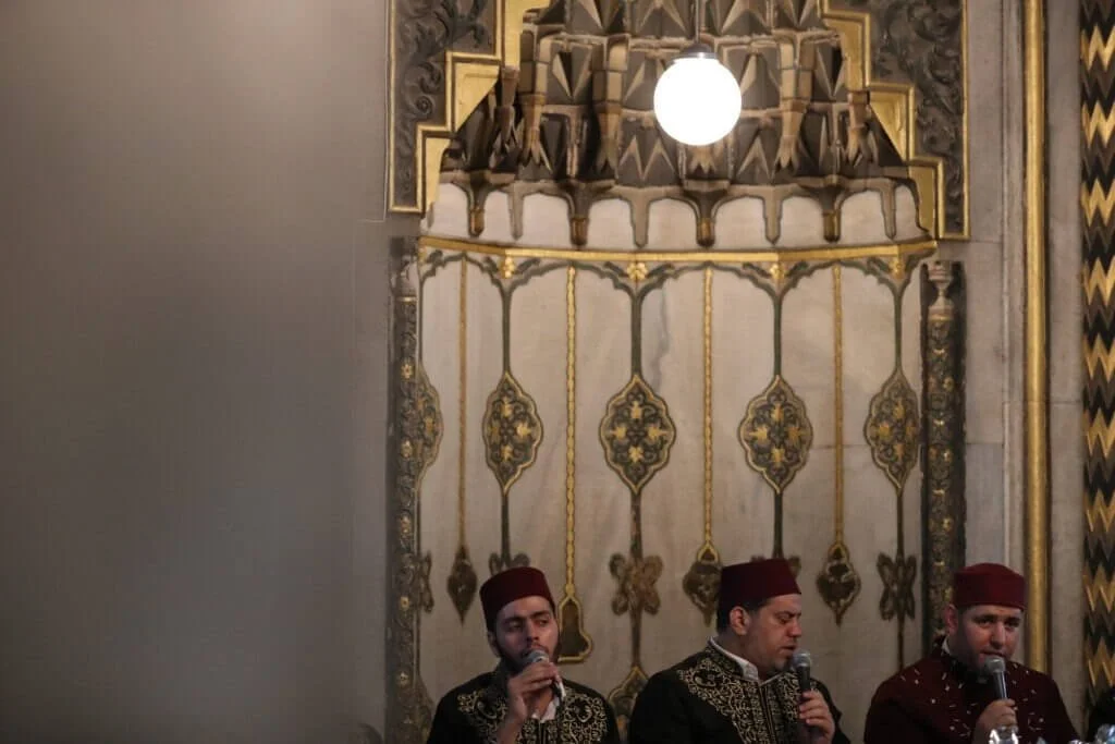 Three men wearing traditional embroidered garments and red caps, sitting and singing into microphones, in a room with ornate gold and gray decor and a hanging white spherical light.