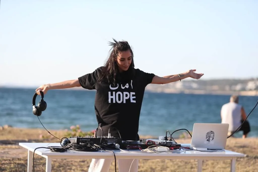 A woman with long dark hair standing outdoors near the ocean, wearing a black T-shirt with 'HOPE' written on it, and holding headphones in one hand. A laptop and DJ equipment are set up on a white table in front of her, with a person in the background facing the water.