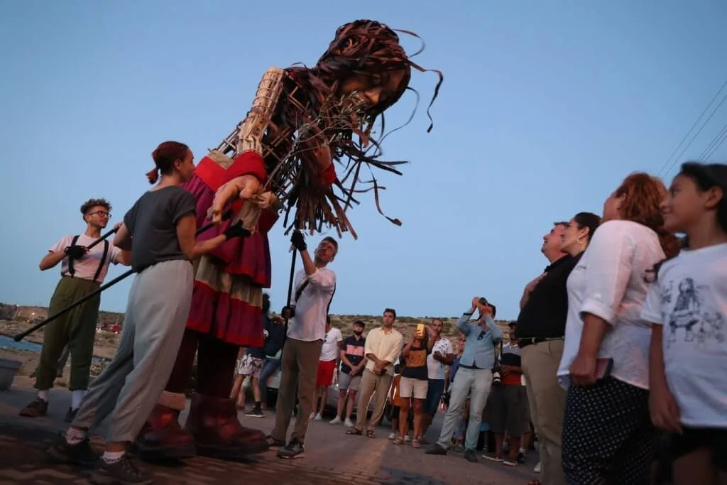 Group of people watching a giant puppet of a person with long hair being carried during an outdoor event at dusk.