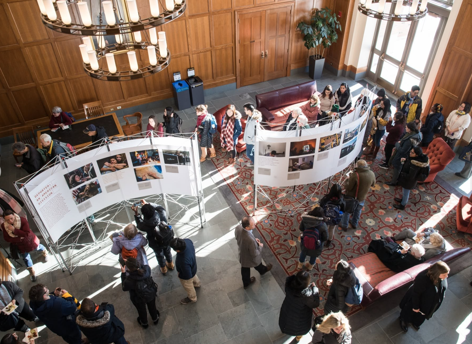 People attending an art exhibit inside a building with wood-paneled walls, large windows, and two overhead chandeliers, viewing photographs displayed on curved white panels.