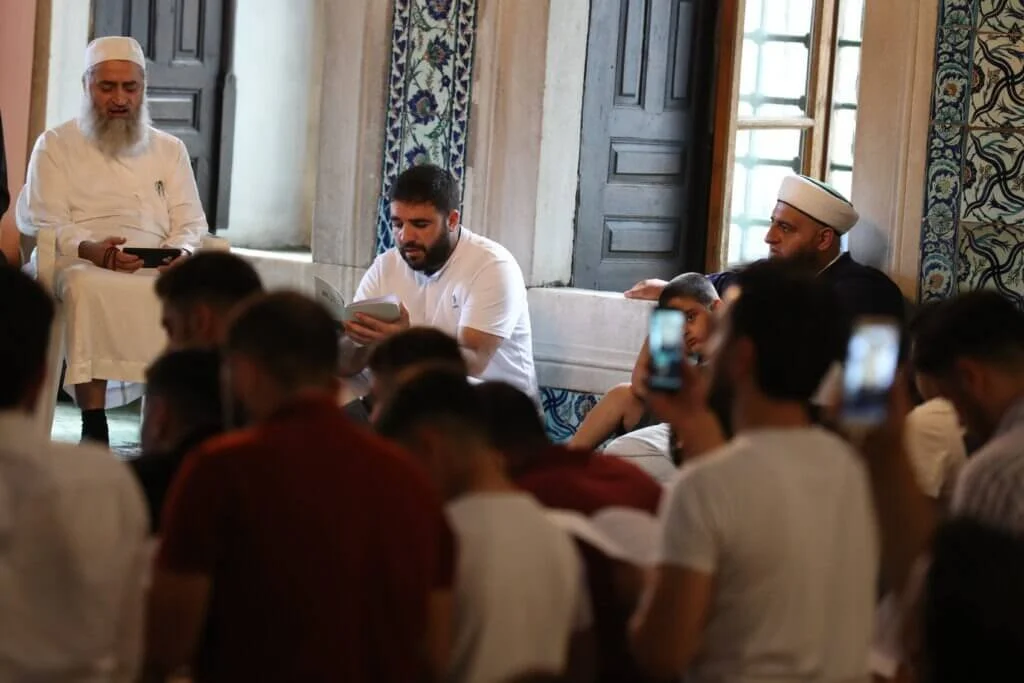 Group of people gathered indoors, sitting and listening to two men at the front, one in traditional Islamic clothing and the other in casual white attire, with some attendees recording the event on their phones.