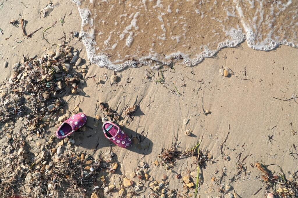 Pink children's shoes with white and yellow flowers on sand at the beach near seaweed and small shells.