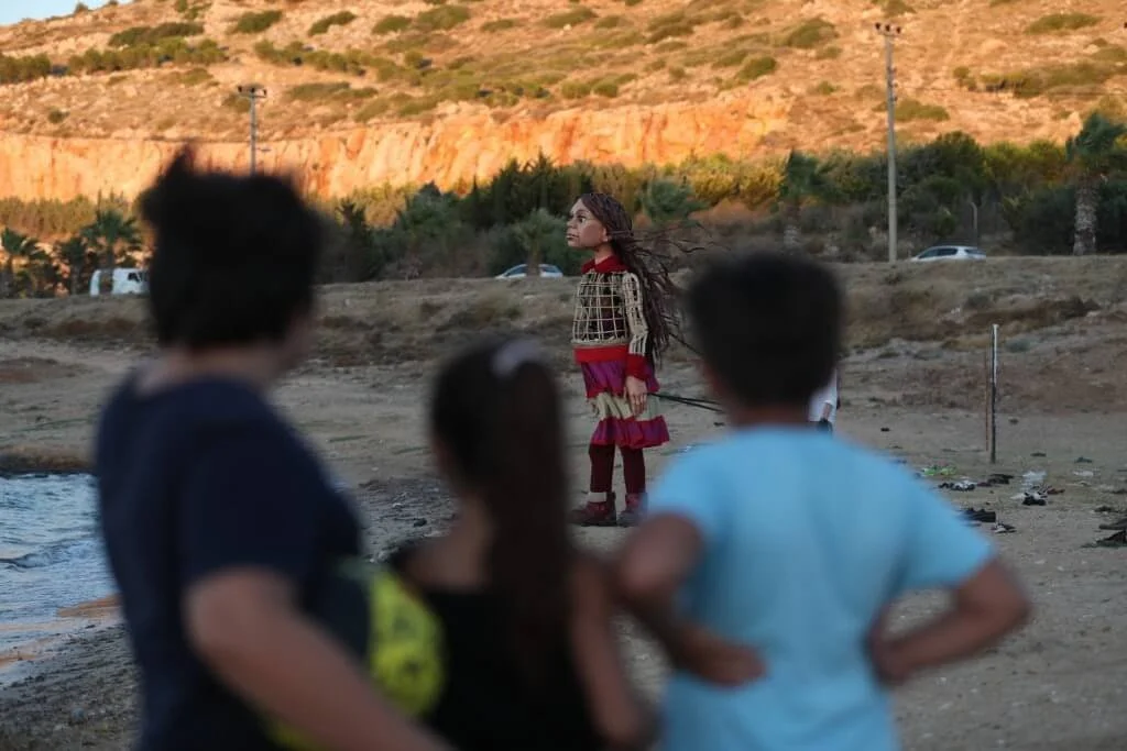 A woman with long, braided hair stands on a sandy beach, facing right, in front of a group of people with blurred faces in the foreground. The background features a hillside with trees, a few cars, and electrical poles.