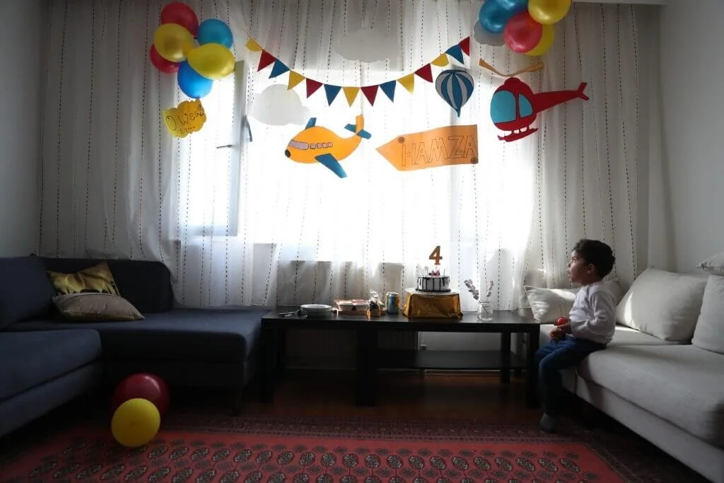 Child sitting on a white sofa at a birthday celebration with colorful balloons, banners, and themed decorations, including a cake on the table celebrating a fourth birthday.