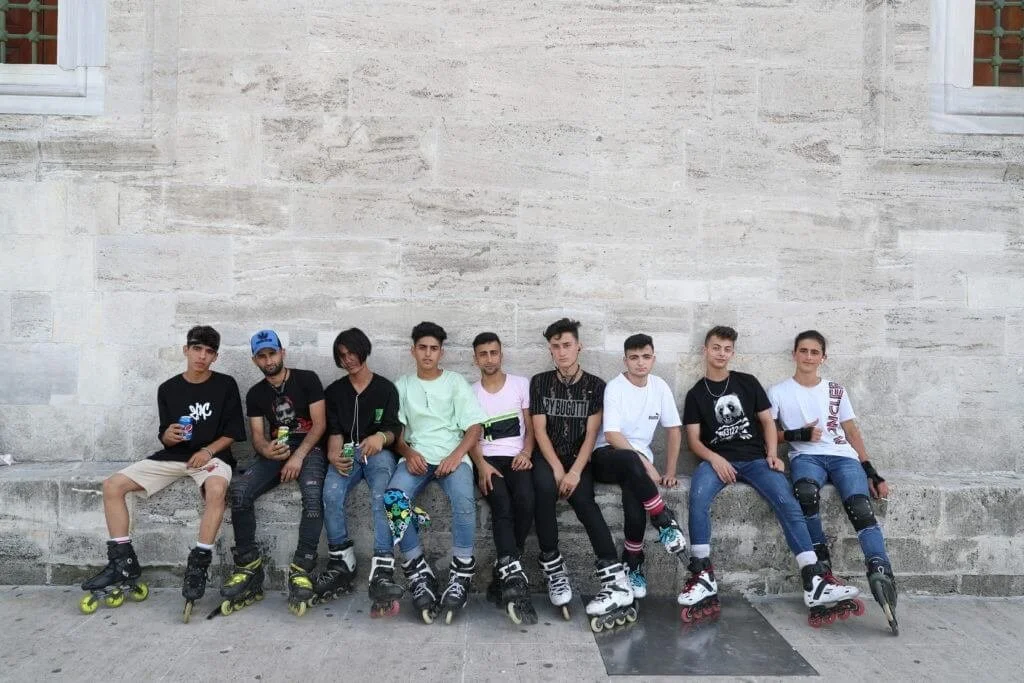 A group of nine young men sitting on a stone ledge, wearing rollerblades and casual clothing, against a large stone wall.