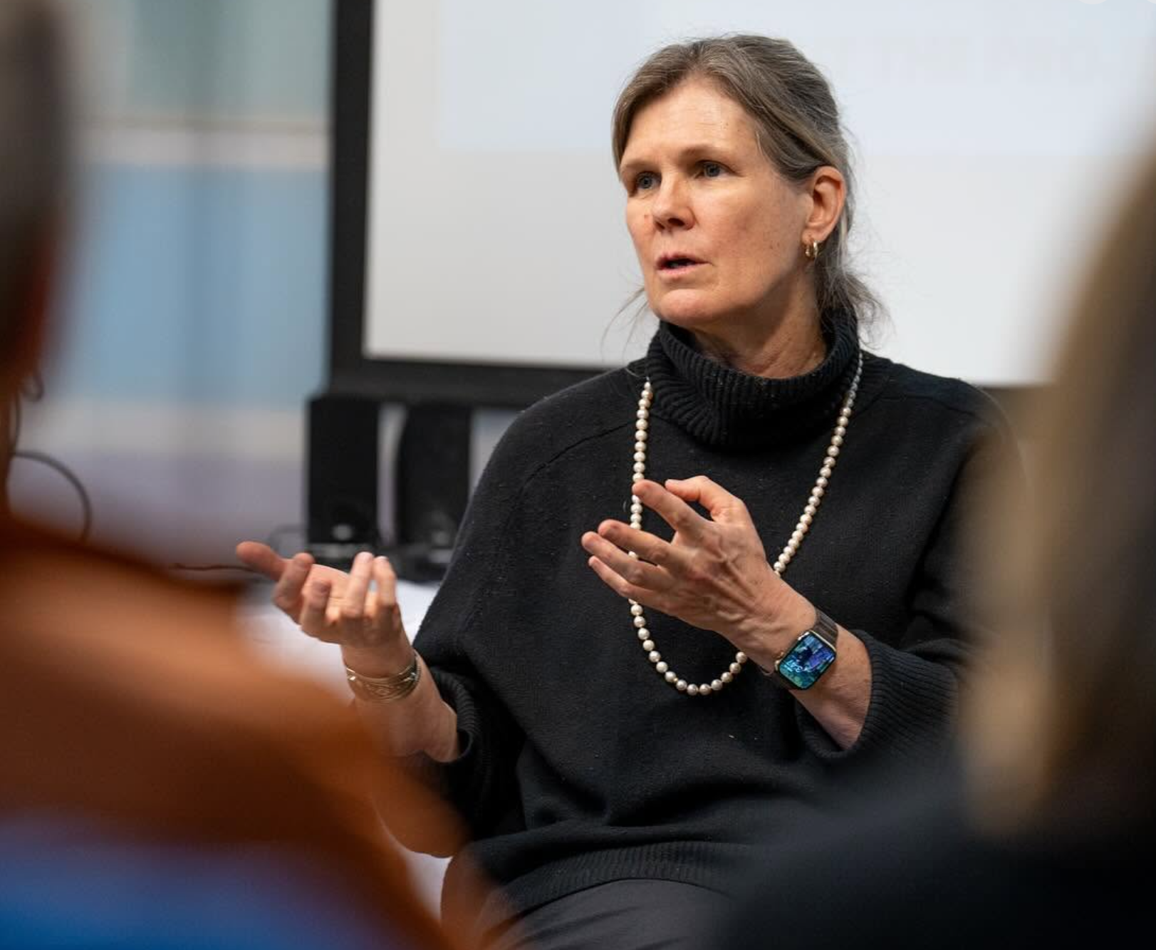 A caucasian woman speaking in a discussion, wearing a black turtleneck sweater, pearl necklace, a watch, and earrings, with a computer monitor in the background.