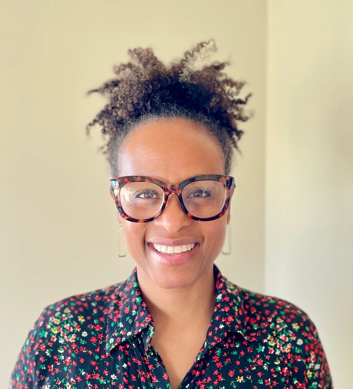 Portrait of a smiling African American woman with curly hair in a bun, wearing large tortoiseshell glasses, a nose piercing, gold earrings, and a floral shirt.