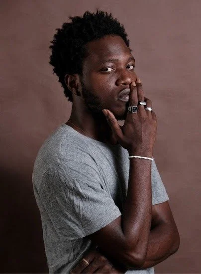 Young African man with curly hair and a beard, wearing a gray t-shirt, posed with his hand near his face, against a plain brown background.