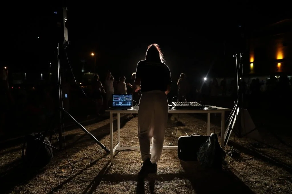 A DJ performs at an outdoor night event with a crowd in the background, illuminated by minimal lighting.