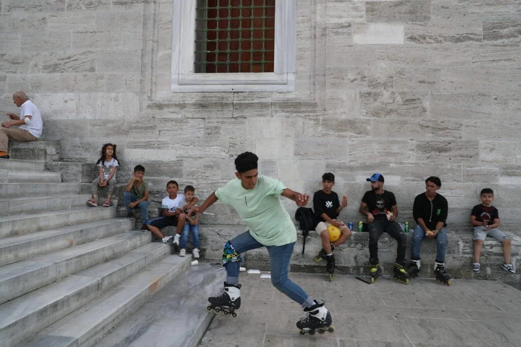 A young man rollerblading down stairs in front of a group of children and teenagers sitting on a ledge against a stone wall, with some watching and others looking away.