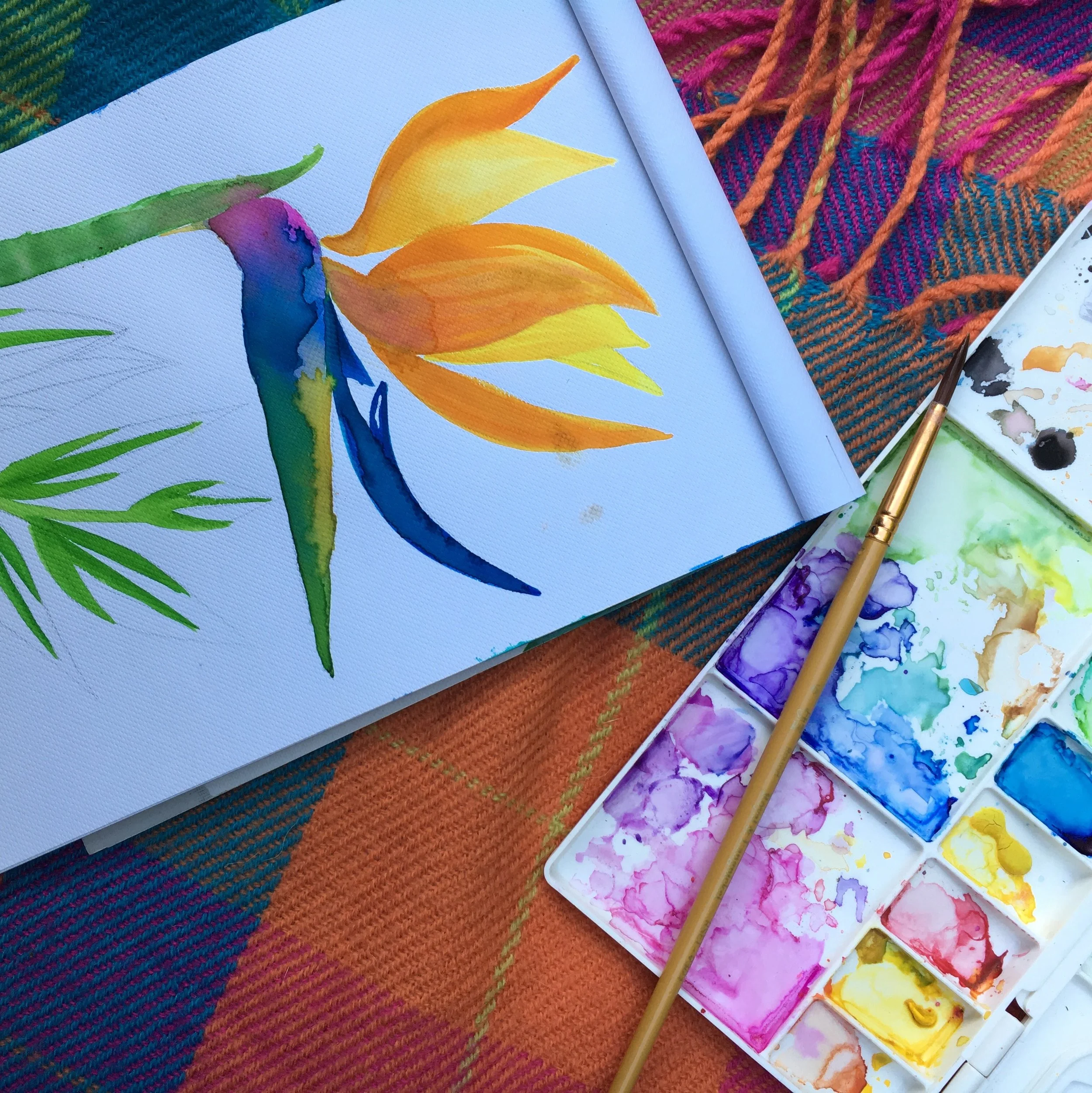 Watercolor painting of a colorful hummingbird with orange and yellow flowers, set on a multicolored woven fabric background.